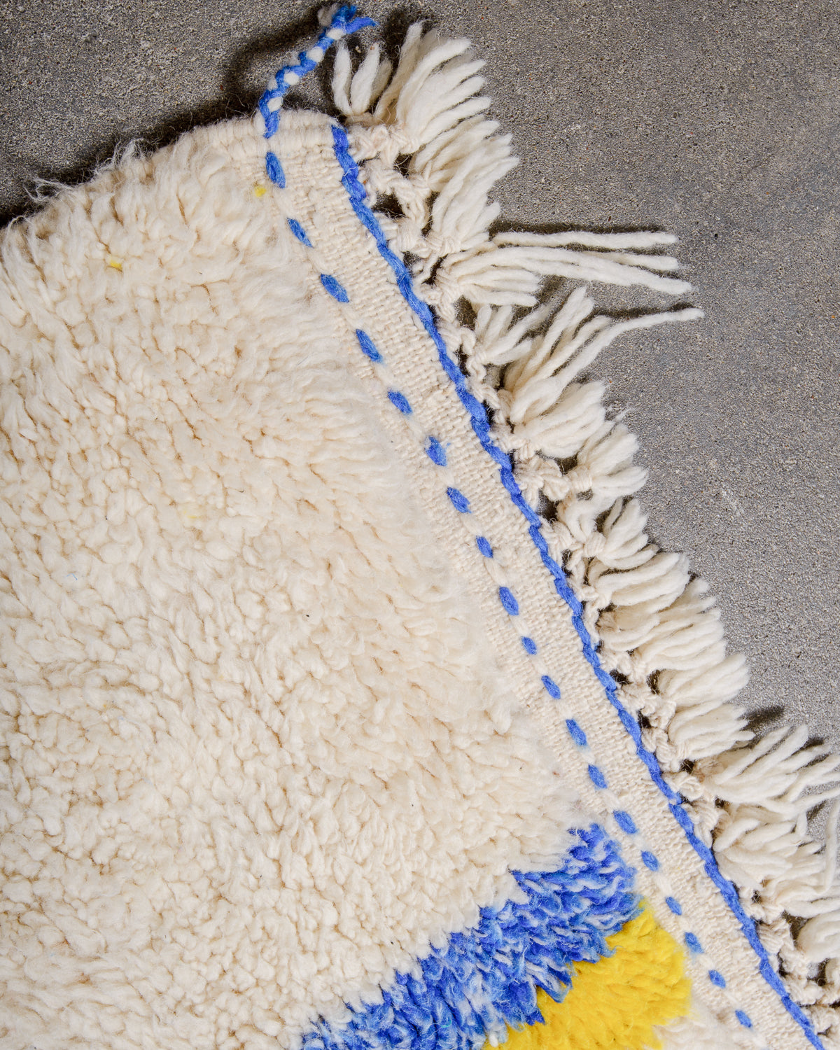 Corner of cream berber rug that is finished with tightly woven ends with blue details and natural wool tassels across the ends..